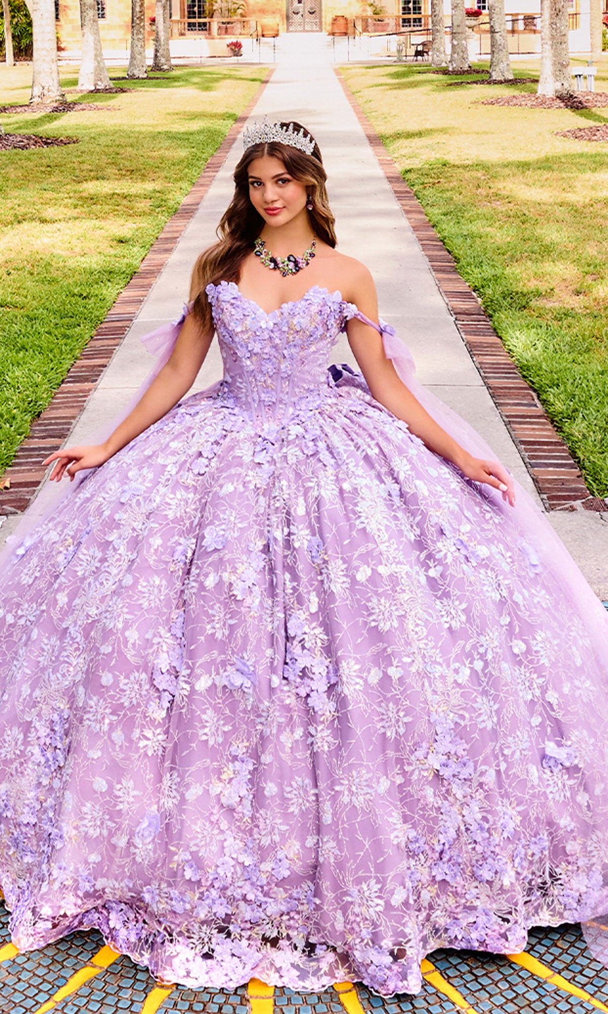 A young woman stands outdoors in the Princesa PR30135 3D Floral Quinceanera Dress, featuring lavender hues, 3D flowers, and a corset bodice, her sparkling tiara catching the sunlight as she faces the camera.